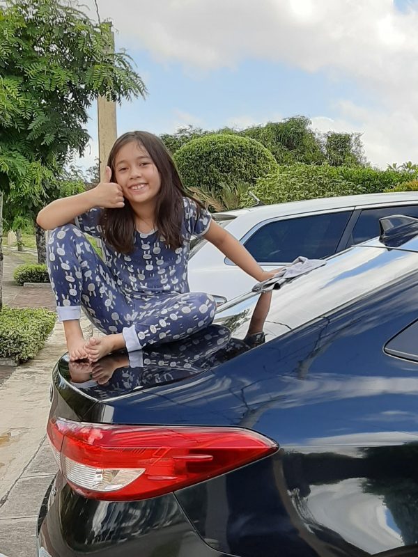 Alba sitting cross-legged on the trunk of a parked black sedan, barefoot and wearing patterned pajamas, giving a thumbs-up and smiling at the camera; cleaning cloth beside her on the trunk; suburban street with trees and another parked car visible, bright daytime, playful and cheerful mood