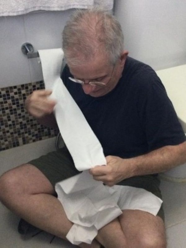 David kneeling on a bathroom floor playing with toddler Alba, who is pulling and wrapping toilet paper from a wall-mounted roll; bathtub and toilet visible nearby, playful and amused atmosphere